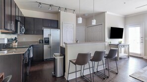 A modern kitchen with dark cabinets, a stainless steel refrigerator, a built-in oven, and a bar area with stools. - Landing | Modern 1BD, Gym (Austin)