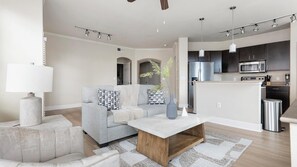 Ein modernes Wohnzimmer mit einer Couch, einem Couchtisch und einer Lampe; eine Küche mit Geräten; und ein Essbereich mit Tisch und Stühlen.