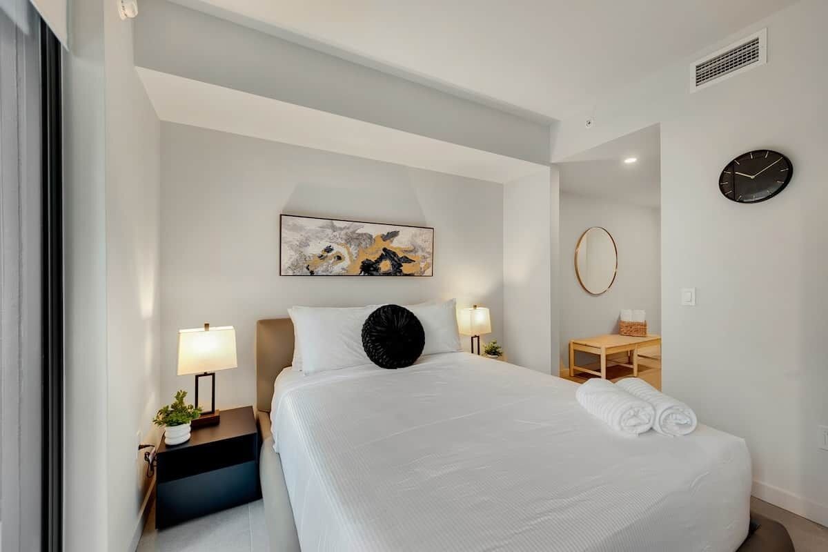 A hotel room with a bed, bedside tables, a desk, a clock, and a painting on the wall.