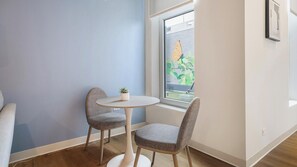 A small room with a round table and two chairs, a potted plant, and a window with a view of a building and greenery. - Landing | Spectacular 1BD in Lincoln Park (Chicago)