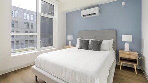 A modern bedroom with a large bed, a window, an air conditioner, and a nightstand with a lamp. - Landing | Spectacular 1BD in Lincoln Park (Chicago)