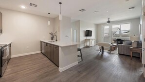 A modern kitchen with a central island, a dining area with a sofa and table, and a living room with a TV. - Landing | Incredible 3BD, Pool, Clubhouse (Goodyear)