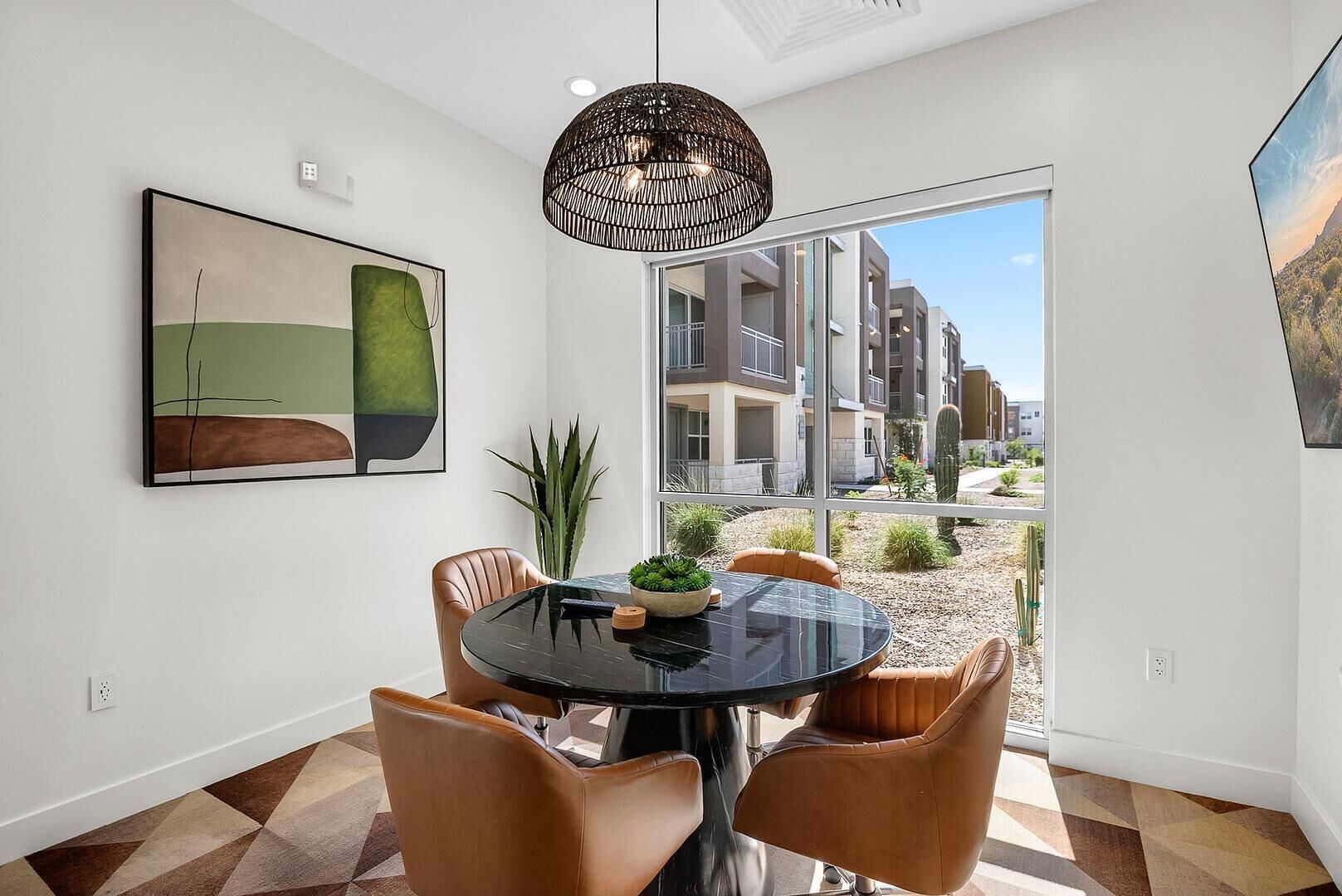 Un coin repas avec une table ronde, des chaises en cuir, une nappe noire, un luminaire suspendu et une grande fenêtre donnant sur des bâtiments et des cactus.