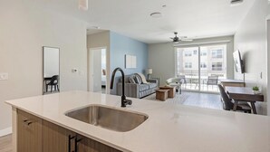 A modern kitchen with a large island, a dining area with a table and chairs, a sofa, and a large window with a view of buildings. - Landing | Modern 2BD, Clubhouse, Pool (Goodyear)