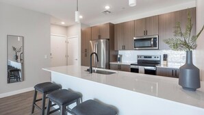 A modern kitchen with stainless steel appliances, a large island with bar stools, and wooden cabinetry. - Landing | Chic 2BD, Clubhouse, Gym (Goodyear)
