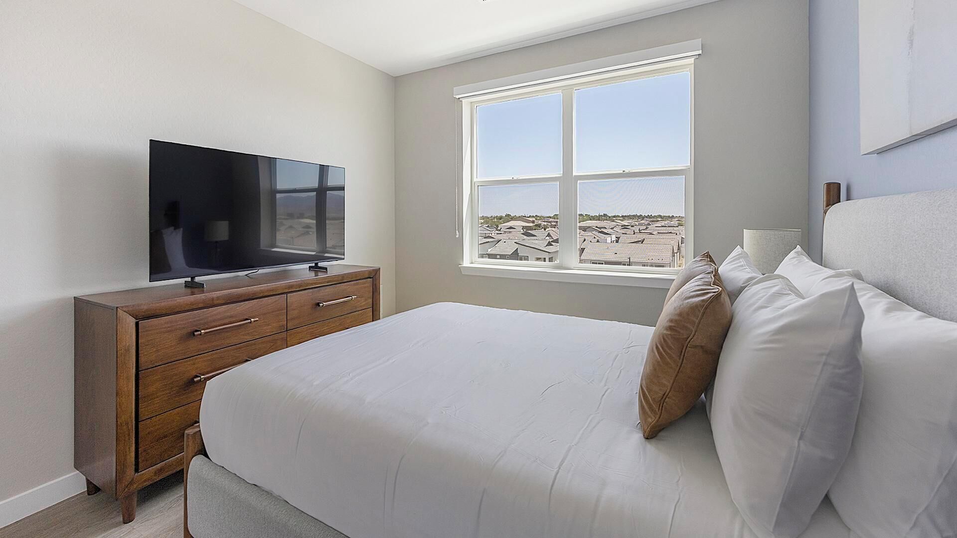 A bedroom with a TV on a wooden dresser, a large bed with white bedding, and a window with a view of buildings.