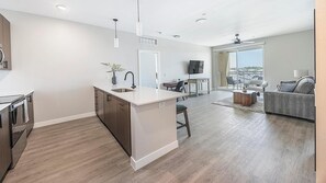 A modern kitchen with a central island, a dining area with a table and chairs, a living room with a sofa, and a balcony with a view.