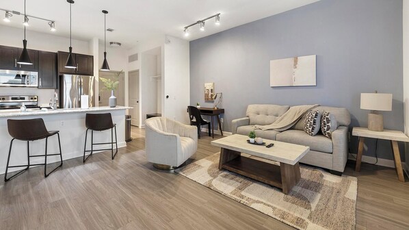 A modern living room with a sofa, coffee table, and chairs. There is a kitchen area with a refrigerator and stove. - Landing | Stunning 1BD, Gym, Game Room (Fort Worth)