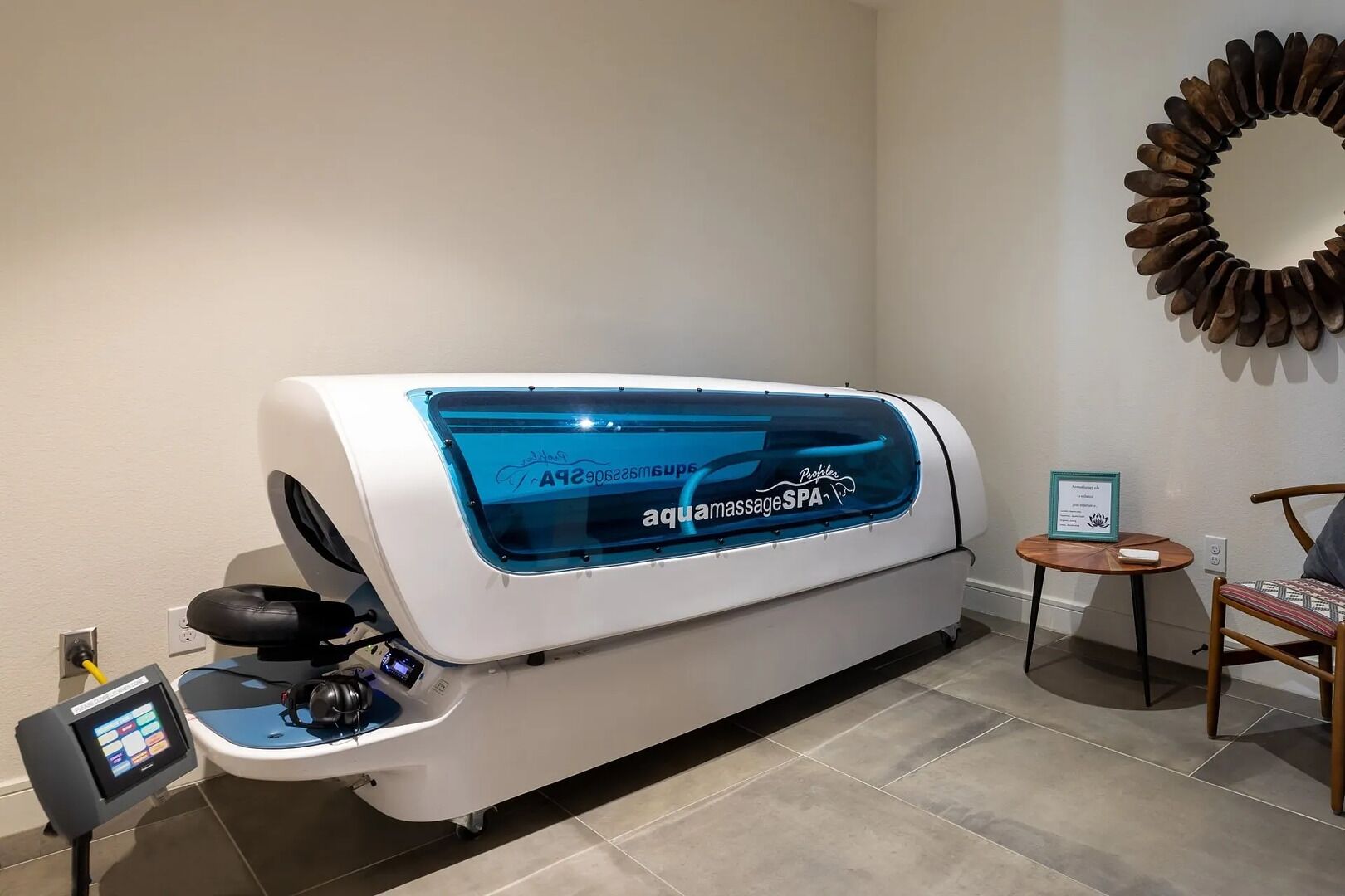 A hydrotherapy machine in a spa room with a control panel, a wooden chair, and a small table with a certificate.