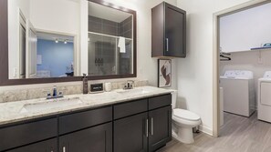A modern bathroom with a double sink, dark cabinets, a toilet, and a shower area. - Landing | Stunning 1BD, Gym, Game Room (Fort Worth)