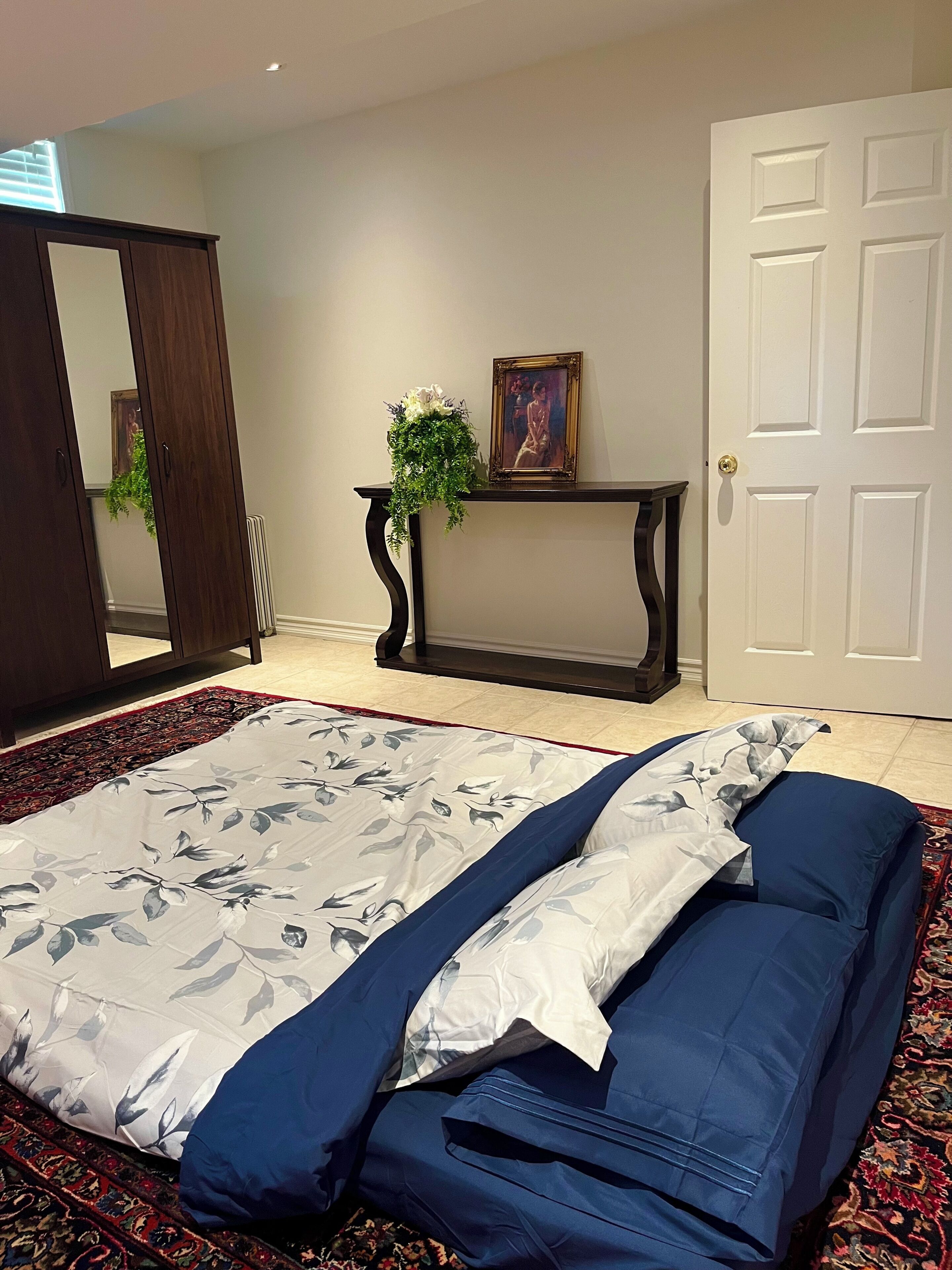 Un dormitorio con una cama, un tocador, una consola con una planta, un cuadro enmarcado y una puerta.
