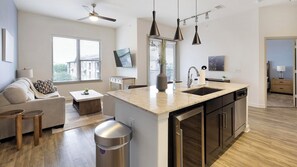 Eine moderne Küche mit einer zentralen Kochinsel, ein Essbereich mit Sofa und Couchtisch sowie ein Wohnzimmer mit Fernseher.