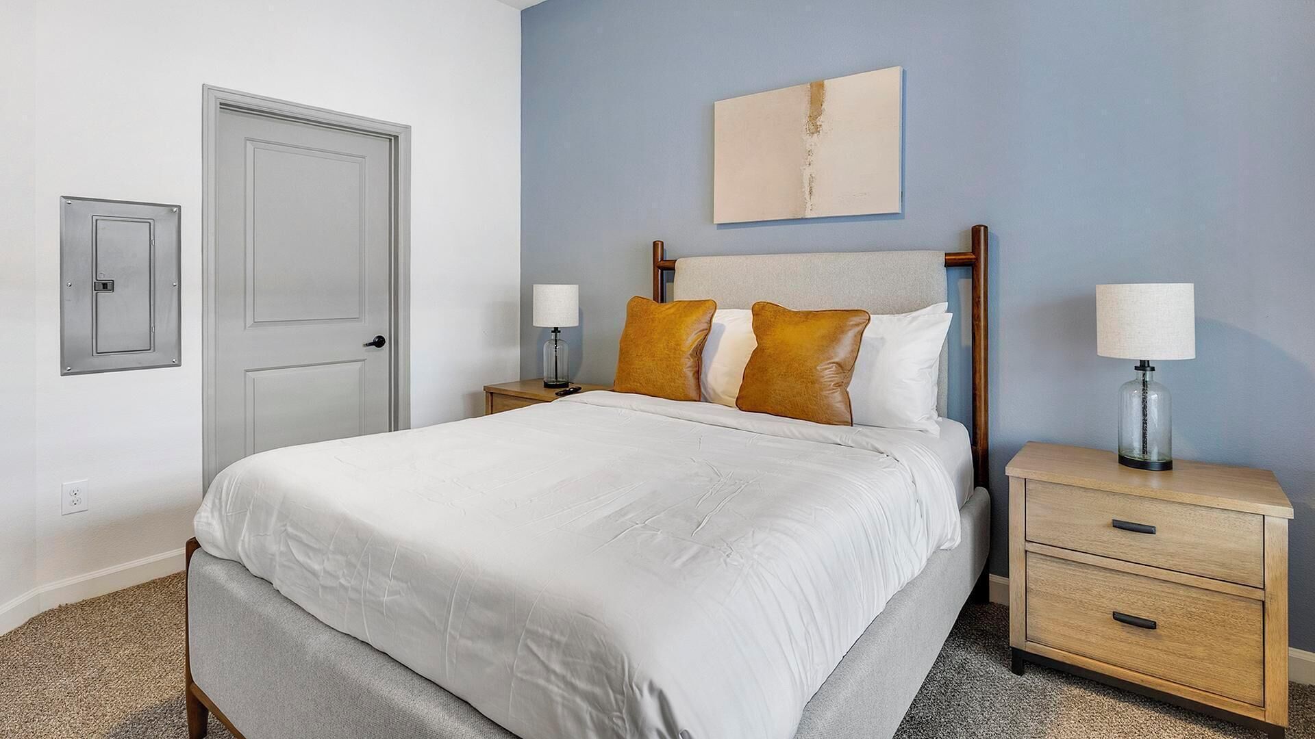 A neatly made bed with a white duvet, two bedside tables with lamps, a wooden headboard, a door, and a wall-mounted artwork.