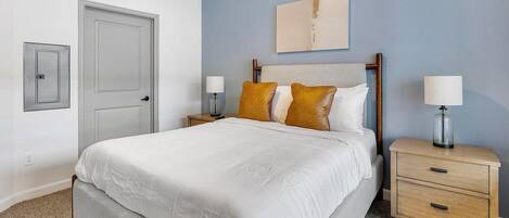 A neatly made bed with a white duvet, two bedside tables with lamps, a wooden headboard, a door, and a wall-mounted artwork.