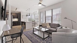 A modern living room with a sofa, coffee table, and a dining area with chairs. - Landing | Amazing 2BD, Gym, Game Room (Fort Worth)