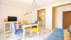 A living room with a sofa, a dining table with chairs, a TV on a cabinet, and a kitchen in the background. - Casa Floriana Seaview - Panoramic Terrace in Capri (Capri)