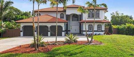 A two-story house with a red-tiled roof, multiple garage doors, and a well-maintained lawn with palm trees.