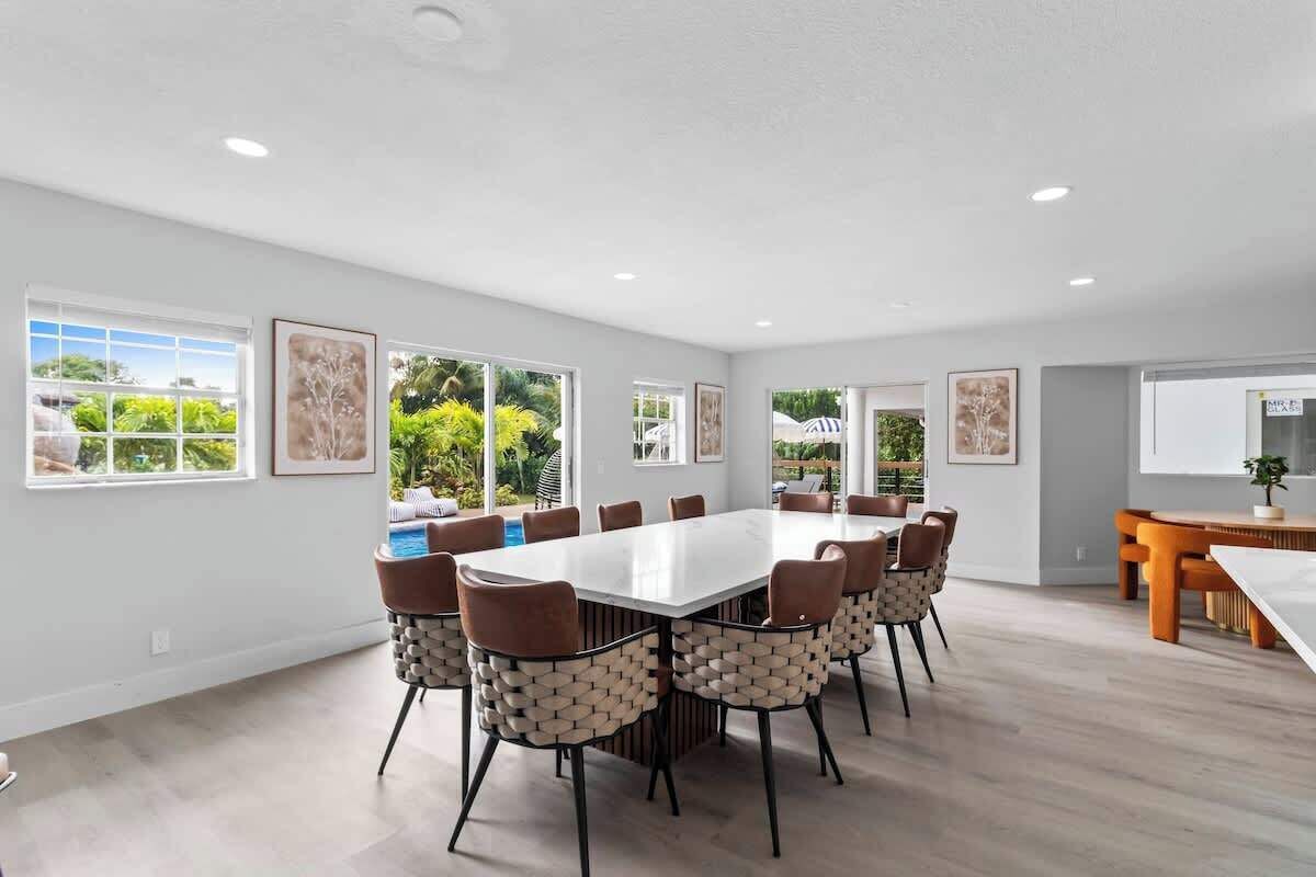 A spacious room with a large white table surrounded by chairs, large windows with views of greenery, and a small wooden table with a plant.