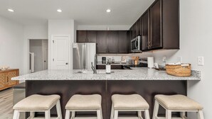 A modern kitchen with dark cabinets, a granite countertop, a stainless steel refrigerator, a microwave, and a built-in oven. There are three bar stools under the island. - Fall Savings - House w/ Pool, indoor games & only 5 miles to Navarre Beach (Navarre)
