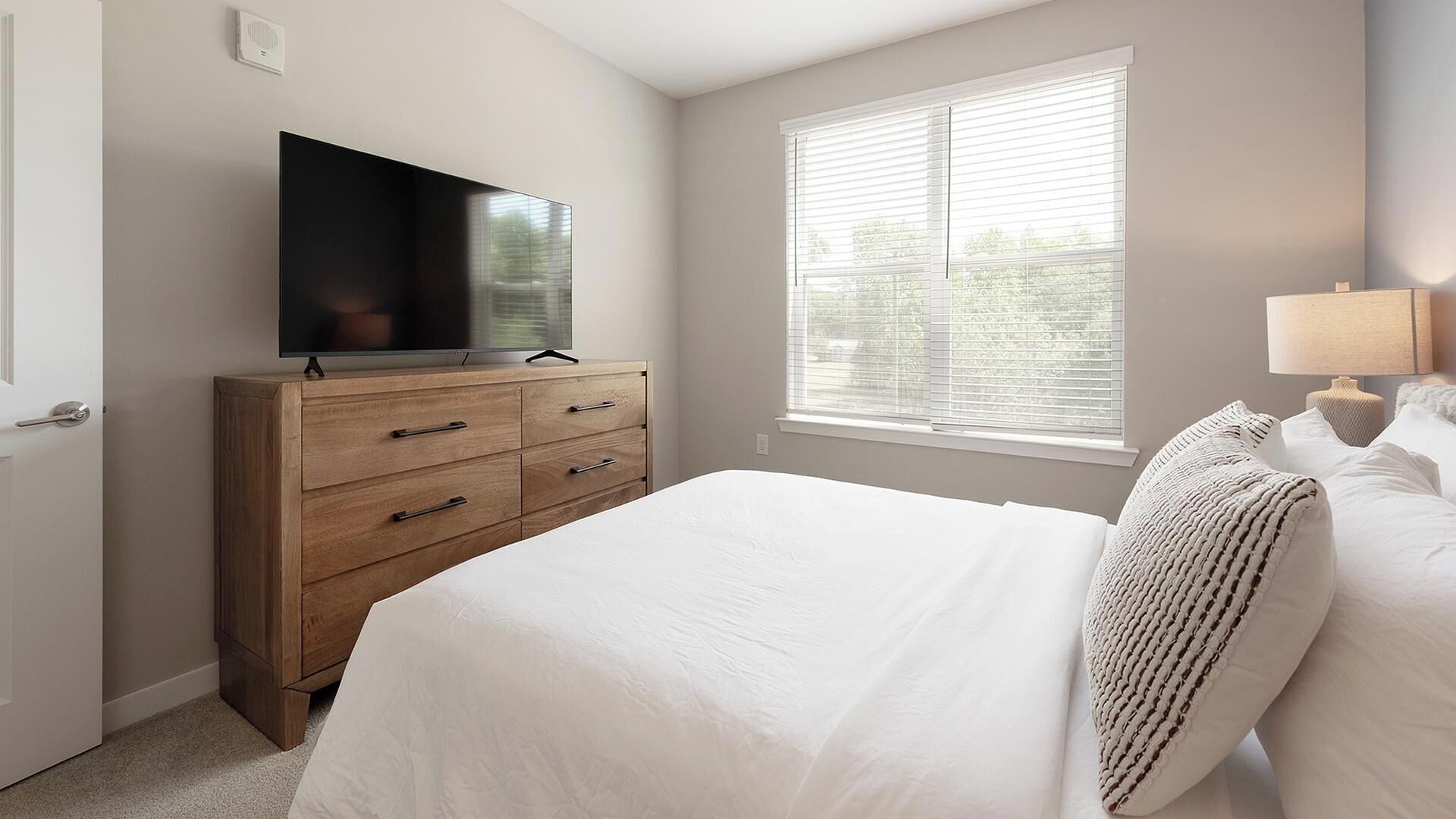 Un dormitorio con cama, televisión sobre un tocador de madera, ventana con persianas y lámpara de noche.