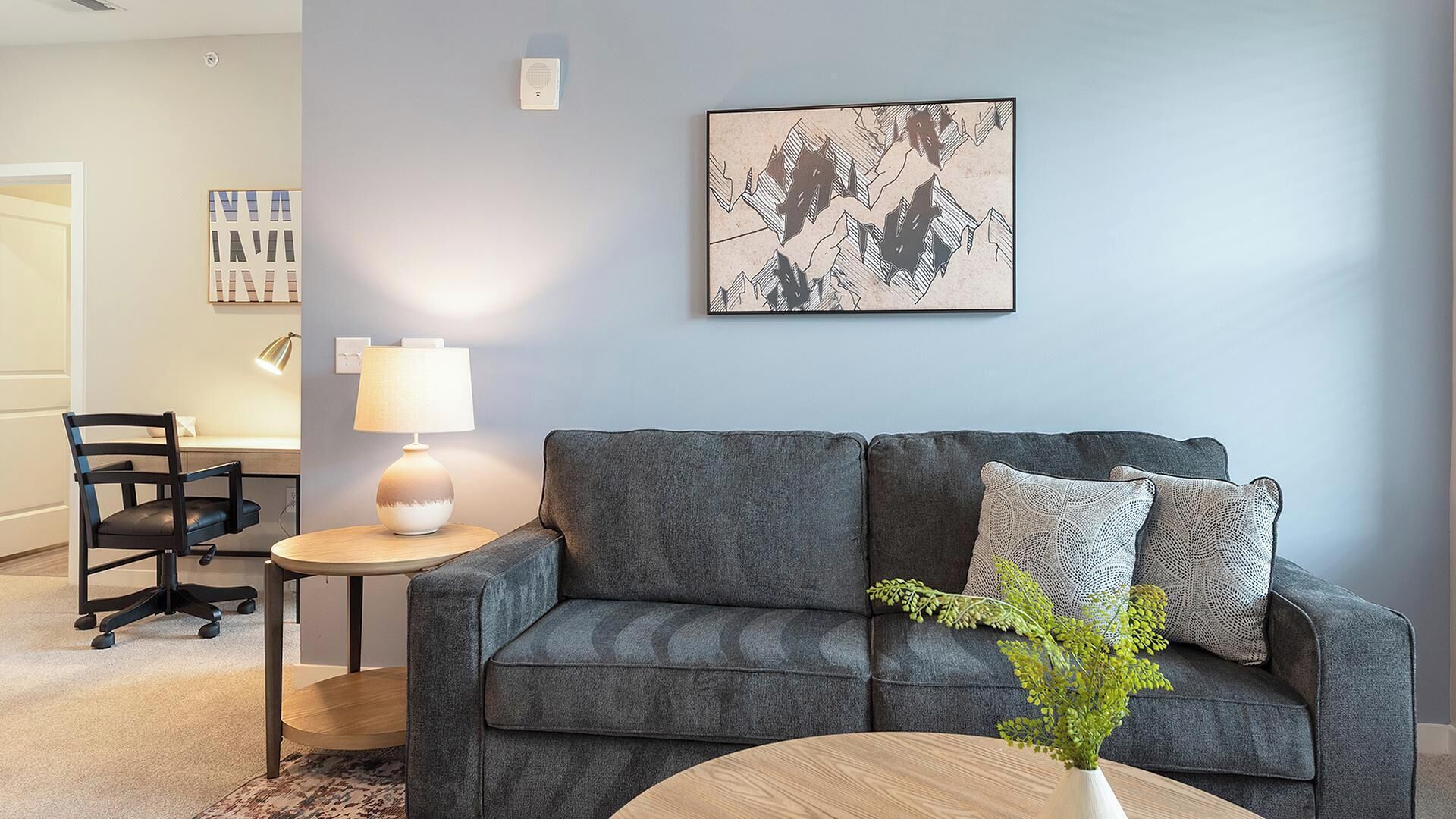 Una sala moderna con un sofá gris, una mesita redonda con una lámpara, una silla y una obra de arte abstracto enmarcada en la pared.