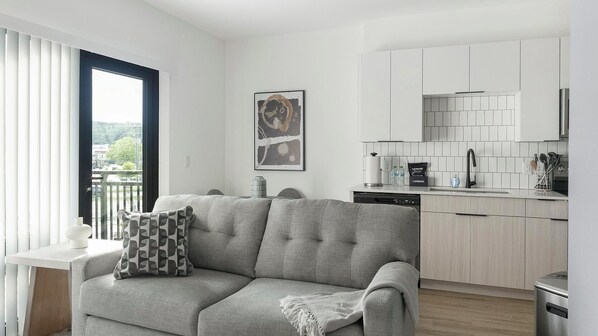 Eine moderne Wohnung mit grauem Sofa, einer Küche mit weißen Schränken und Blick über die Stadt durch das Fenster.