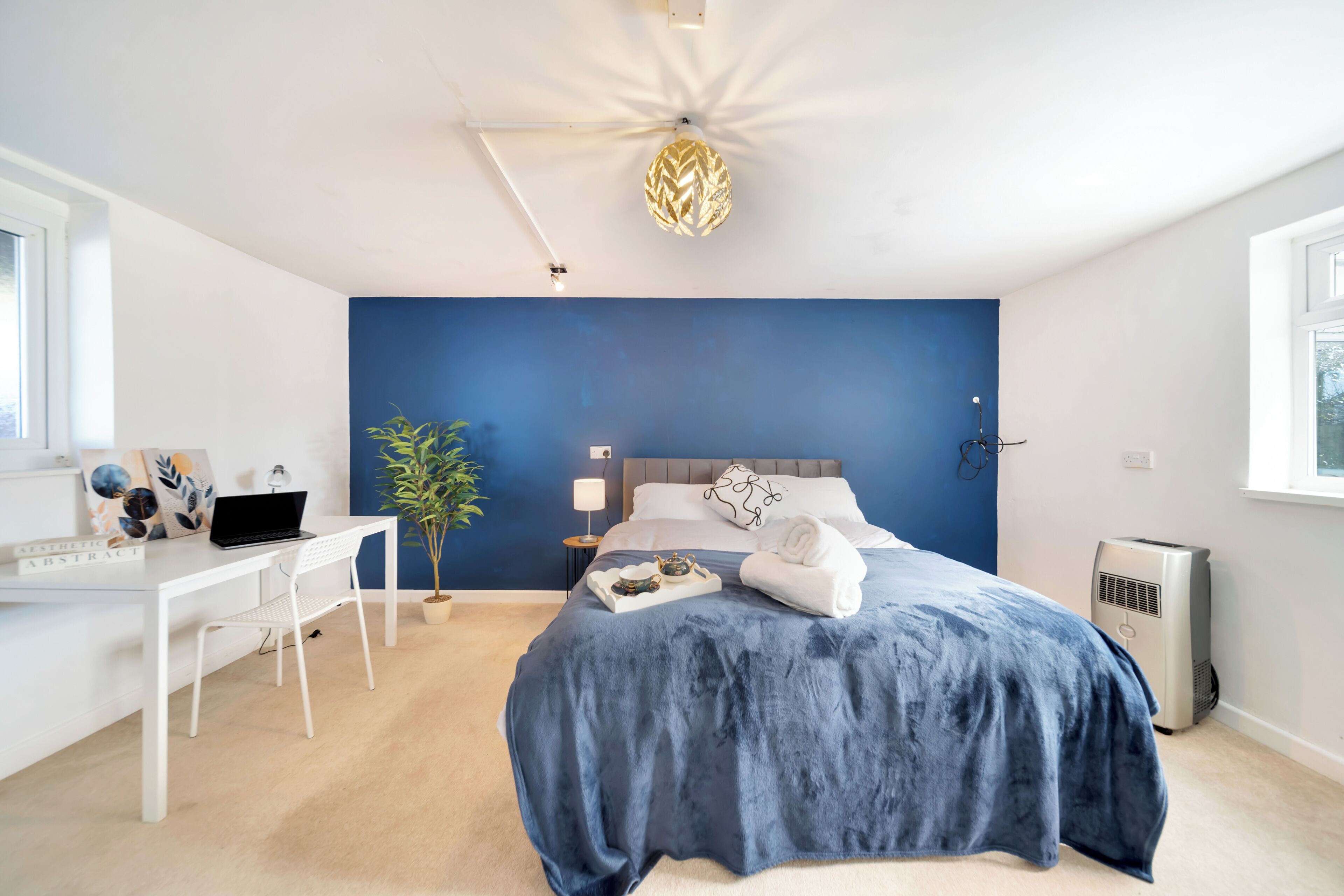 Ein Schlafzimmer mit Bett, Schreibtisch, Laptop, Topfpflanze und Wandleuchte.