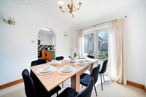 A dining area with a long wooden table set for six, black chairs, a chandelier, and a view through a large window with curtains.