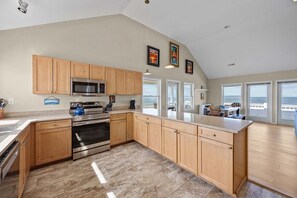 A kitchen with wooden cabinets, stainless steel appliances, and large windows offering ocean views. - 7659 - Sound Garden - Waterfront Avon Retreat | Hot Tub & Sunsets (Avon)