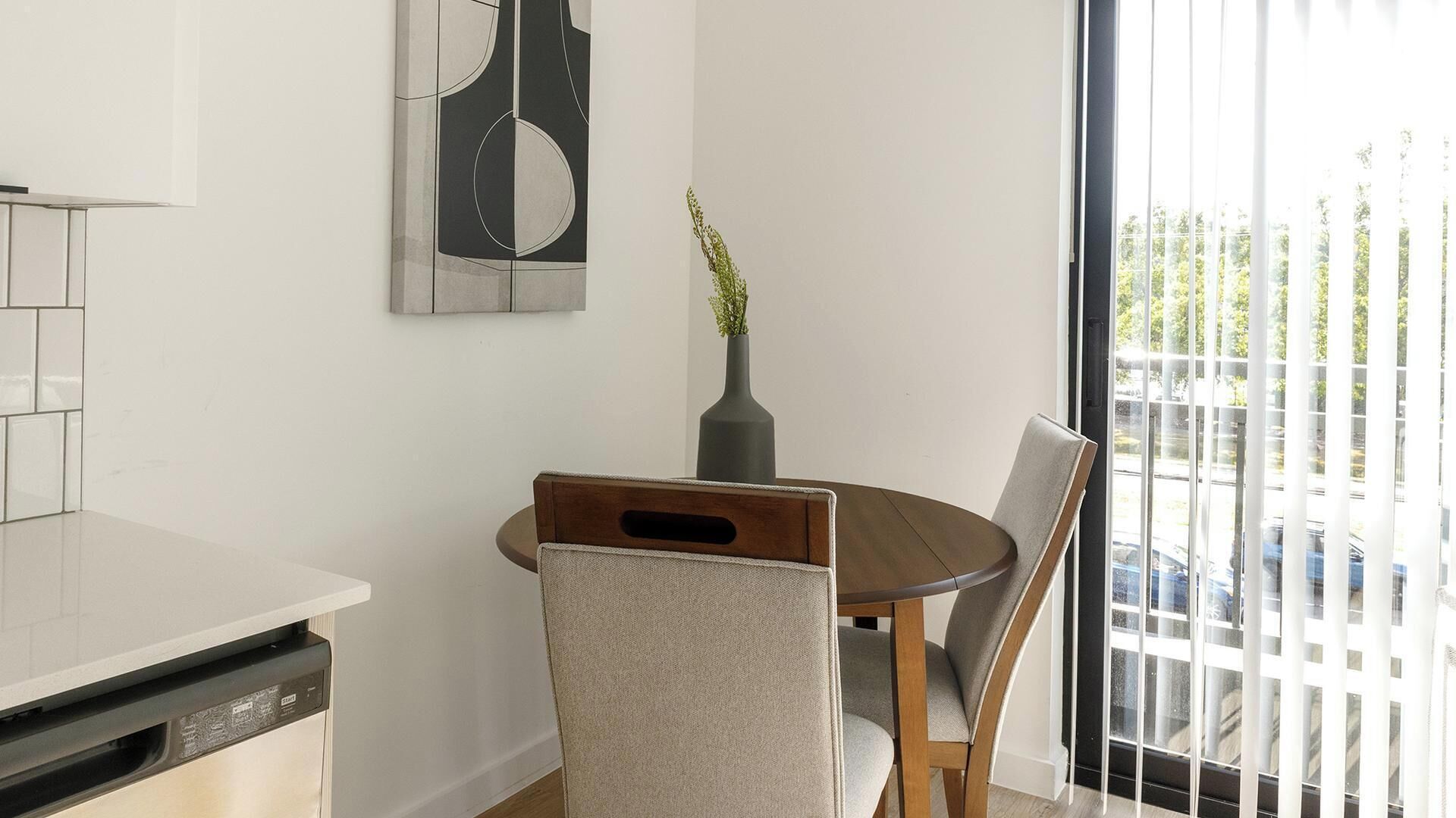 A dining area with a round table, chairs, a vase with greenery, a wall-mounted abstract art piece, and a view through a glass door.