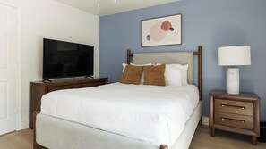 Ein ordentlich eingerichtetes Schlafzimmer mit Bett, Fernseher, Nachttisch mit Lampe und einem Wandbild.