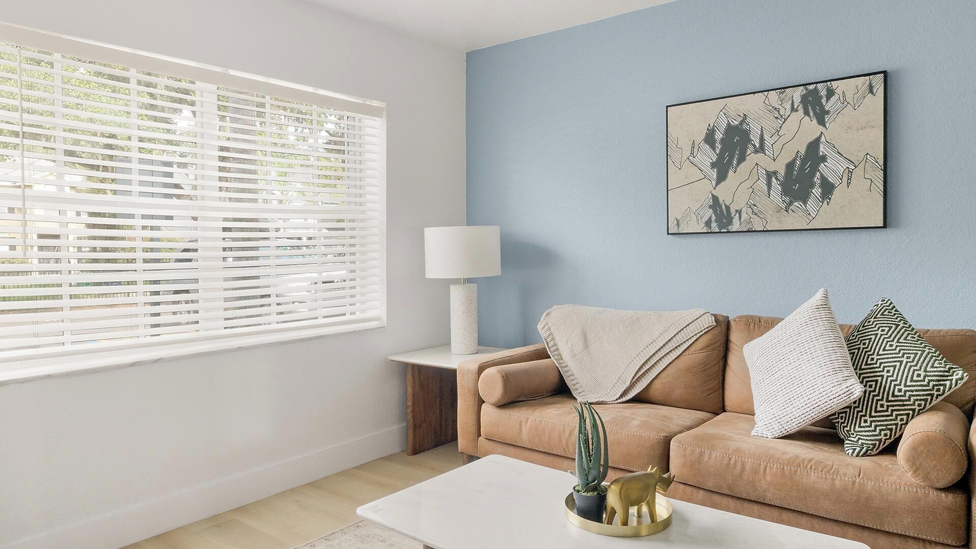 A living room with a beige sofa, a white coffee table, a cactus, a lamp, and a framed artwork on the wall.