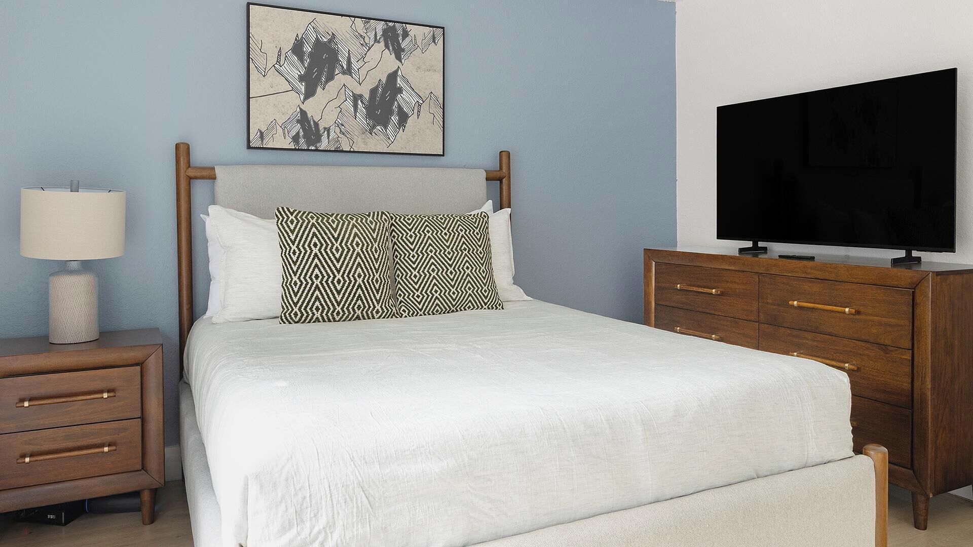 A bedroom with a bed, a nightstand, a dresser, a television, and a wall art piece.