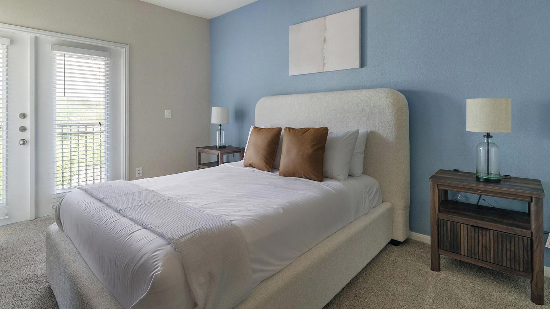A bedroom with a bed, two bedside tables, a door with blinds, and a wall-mounted lamp.