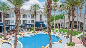 A pool area with palm trees, lounge chairs, and a large building in the background. - Landing | Stunning 2BD, Clubhouse (League City)