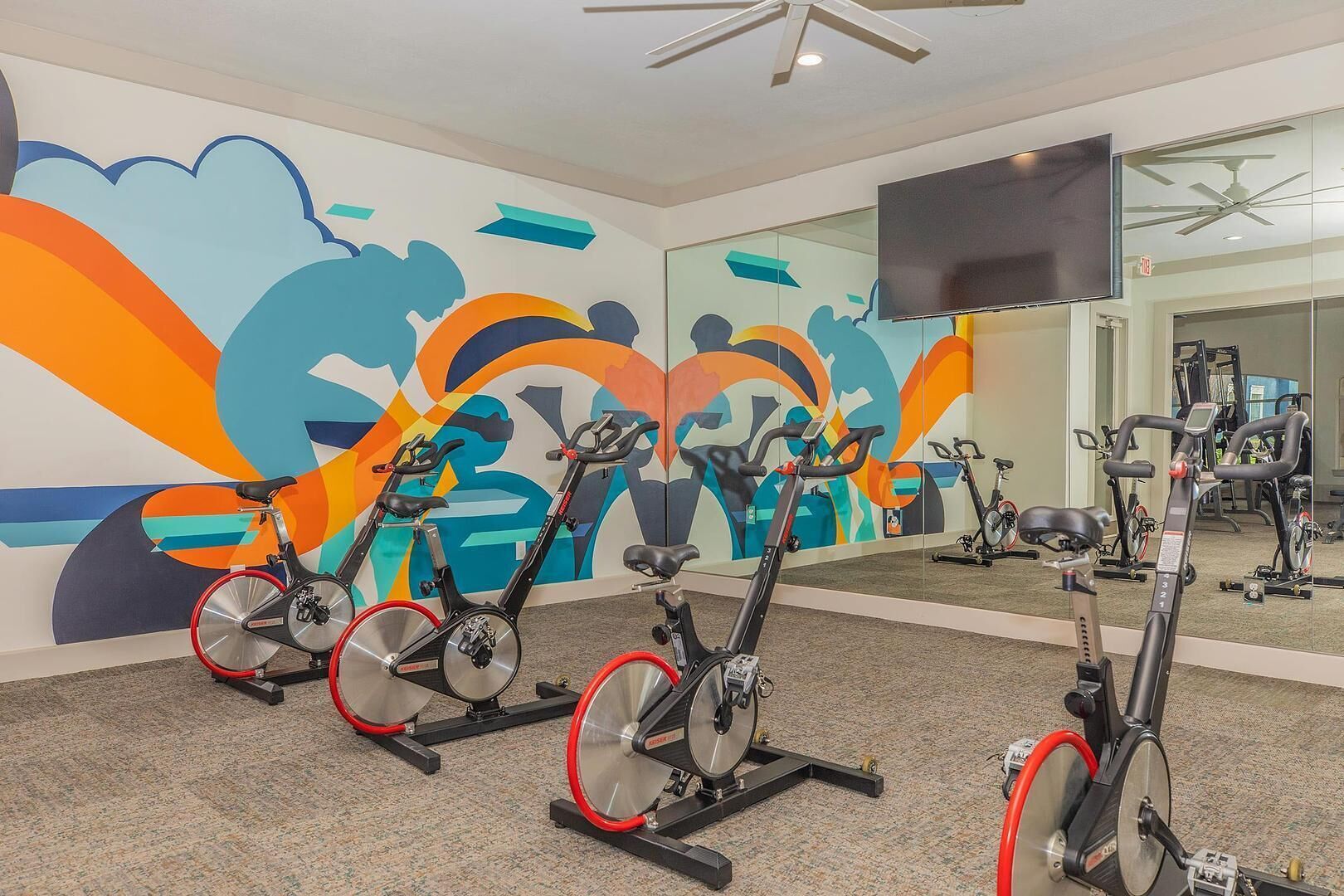 Uma academia com bicicletas ergométricas, um espelho grande e um mural colorido na parede.