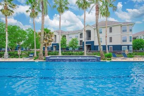 A swimming pool with lounge chairs, palm trees, and a multi-story building with balconies. - Landing | Chic 1BD, Clubhouse (League City)