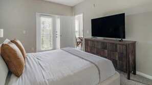 A bedroom with a bed, a TV, a wooden dresser, and a door leading to another room. - Landing | Chic 1BD, Clubhouse (League City)