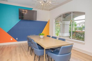 A conference room with a large table, blue chairs, a potted plant, a TV mounted on the wall, and a large window with a view of greenery. - Landing | Incredible 1BD, Clubhouse (League City)