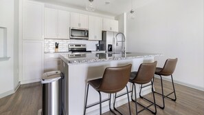 A modern kitchen with white cabinets, a stainless steel island, bar stools, and a trash can. - Landing | Chic 1BD in The Forum (Fort Myers)
