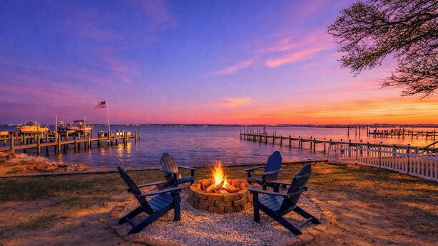 A waterfront outdoor seating area with a fire pit and Adirondack chairs.