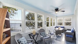 A covered porch with wicker furniture, a ceiling fan, and large windows overlooking a body of water.