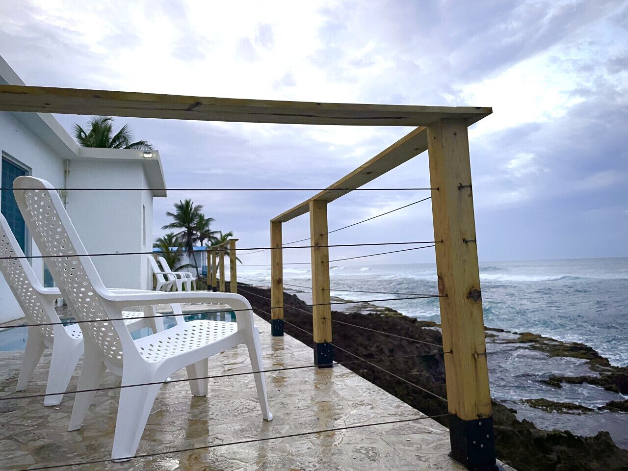 A wooden deck with a railing overlooking the ocean, white plastic chairs, and a building with a blue door in the background.