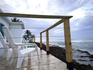 A wooden deck with a railing overlooking the ocean, white plastic chairs, and a building with a blue door in the background.