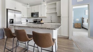 A modern kitchen with white cabinets, stainless steel appliances, and a granite countertop. There are three brown bar stools and a wooden floor.