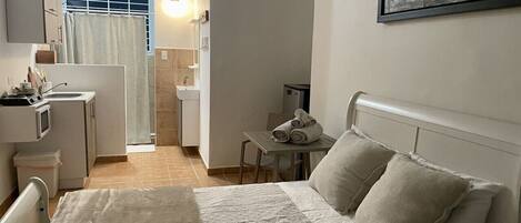 Een slaapkamer met een bed, kussens, een klein tafeltje met handdoeken, een kitchenette en een badkamer die door een open deur zichtbaar is.