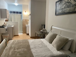 Ein Schlafzimmer mit Bett, Kissen, einem kleinen Tisch mit Handtüchern, einer Küchenzeile und einem durchgegangenes Badezimmer.