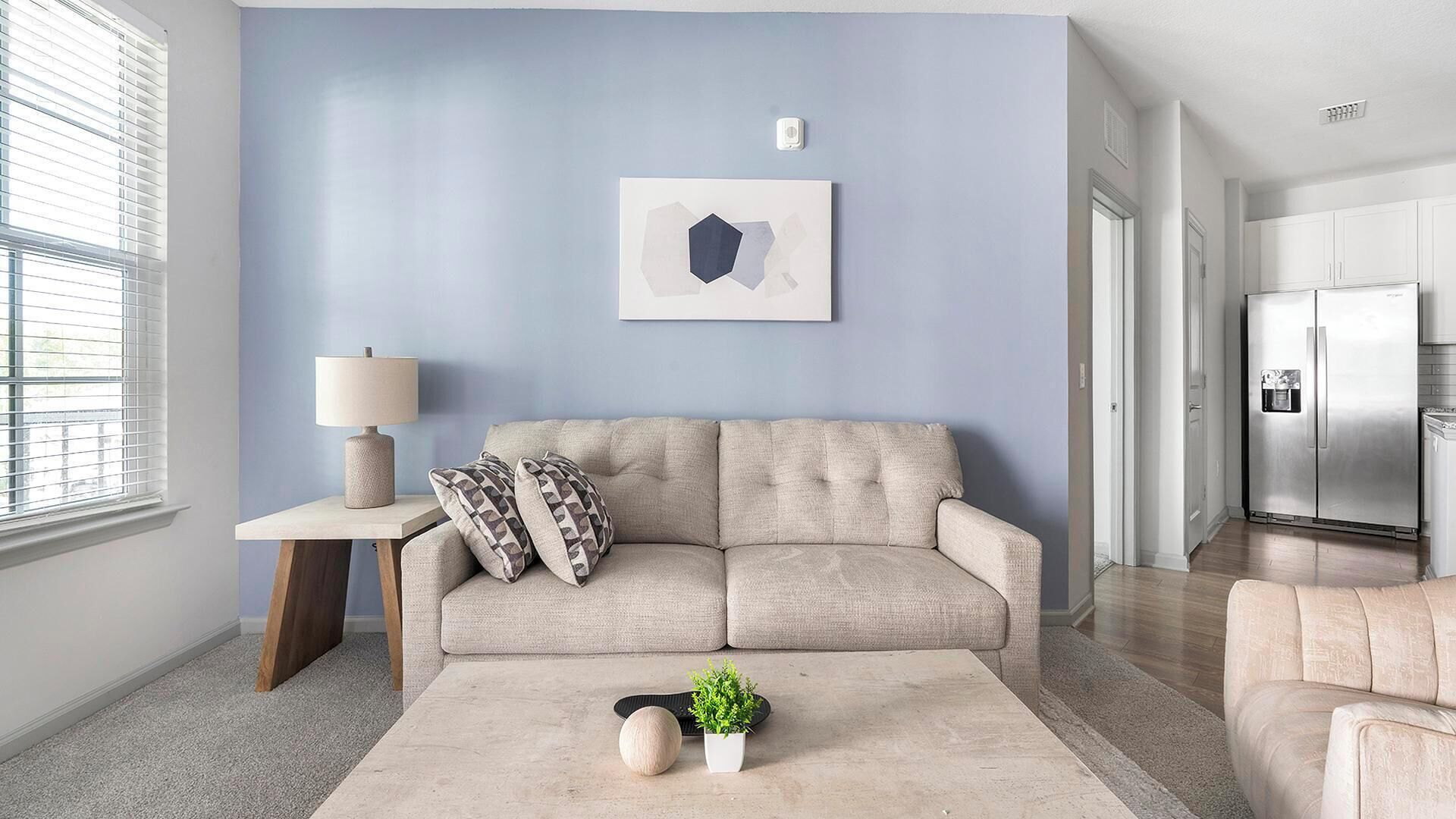 Una sala de estar con un sofá, una mesita de noche con una lámpara, una mesa baja con una planta y una zona de cocina con un refrigerador.