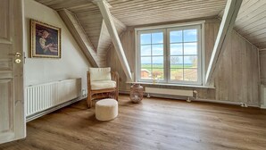Ein Zimmer mit schrägem Dach, einem großen Fenster mit Ausblick, einem Sessel, einem kleinen Hocker und einer Heizung.