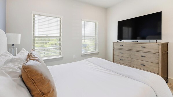 Un dormitorio con una cama, dos ventanas con persianas, un televisor sobre un tocador de madera y una lámpara de noche.